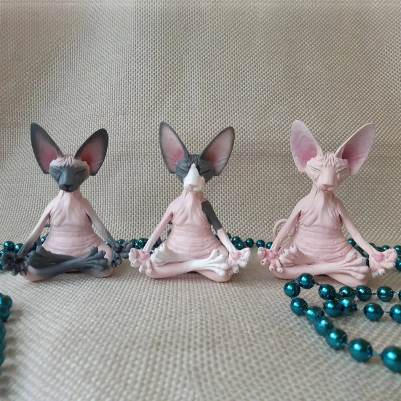 Three Sphynx cat figurines sit in a meditative pose on a textured surface, each with unique patterns and surrounded by turquoise beads.