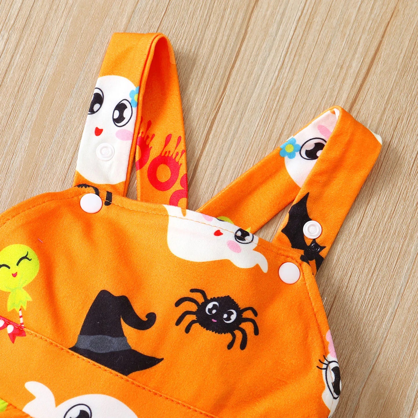 Bright orange fabric with cute Halloween-themed prints, including ghosts, bats, spiders, and a witch hat. The fabric has snap buttons and is set on a wooden surface.