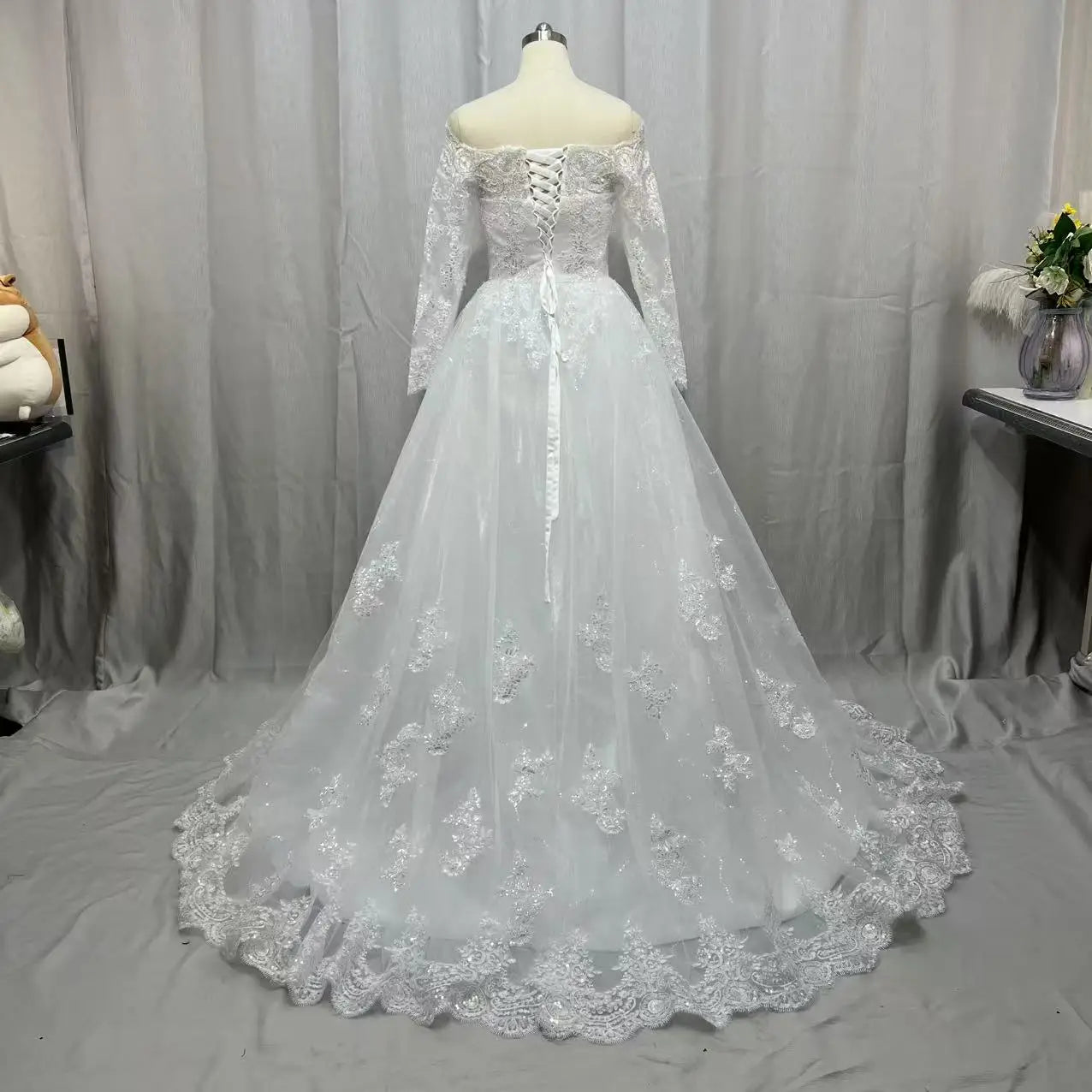 Back view of an off-the-shoulder, long-sleeve wedding dress with intricate lace detailing and a corset back, displayed on a mannequin against a gray backdrop.