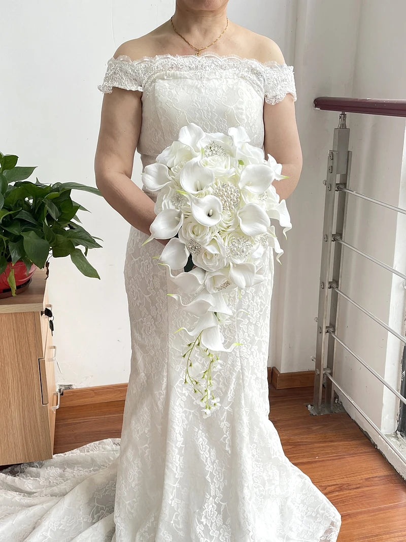A bride in a white lace off-shoulder gown holds a bouquet of white flowers, including calla lilies, standing indoors on a wood floor next to a green potted plant.