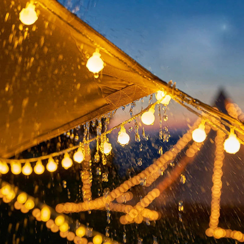 Strings of warm glowing lights hang under a wet canopy during rain, creating a cozy ambiance against a blurred twilight sky.
