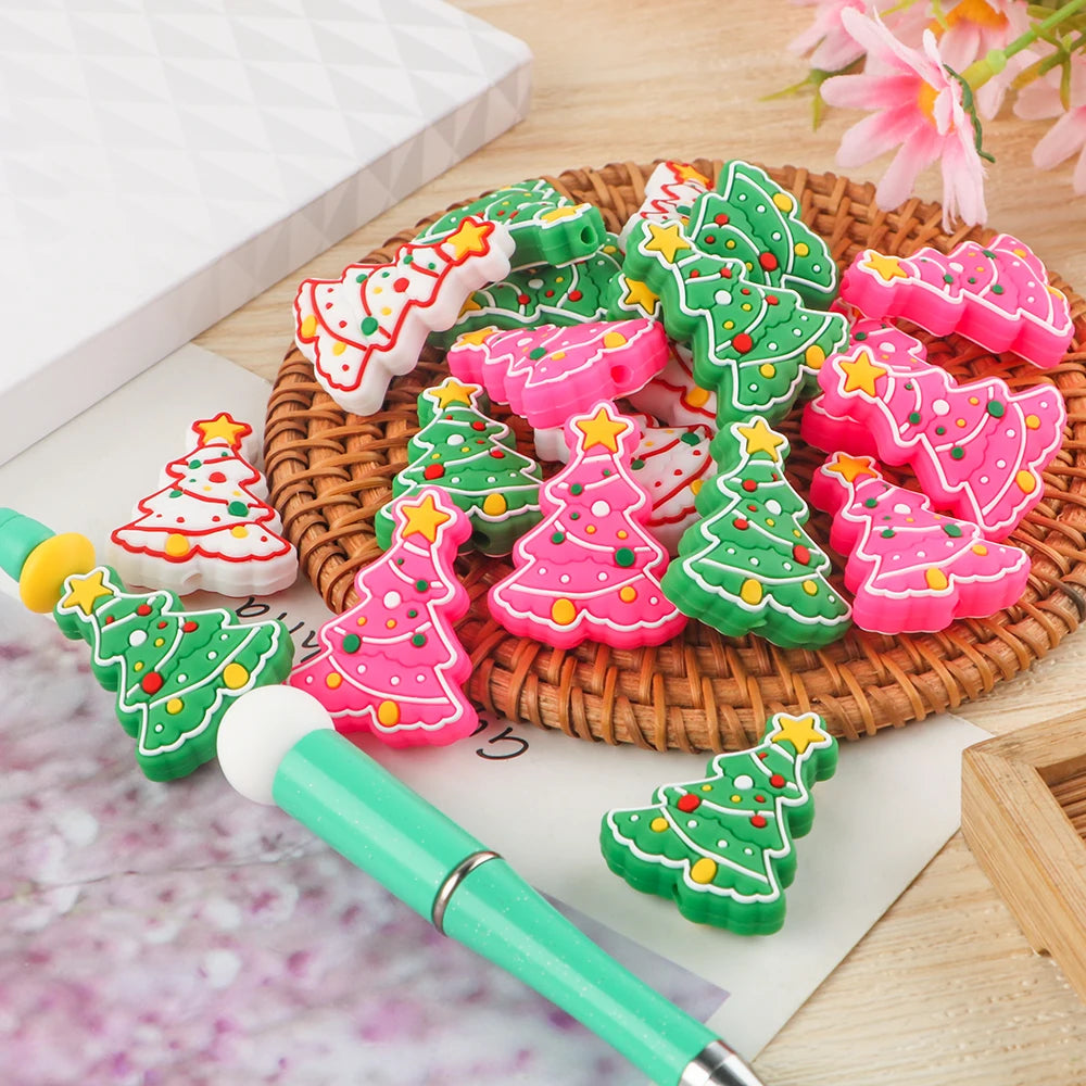 Colorful Christmas tree erasers in pink, green, and white, scattered on a woven mat, with a green pen nearby. A notepad and pink flowers are partially visible.