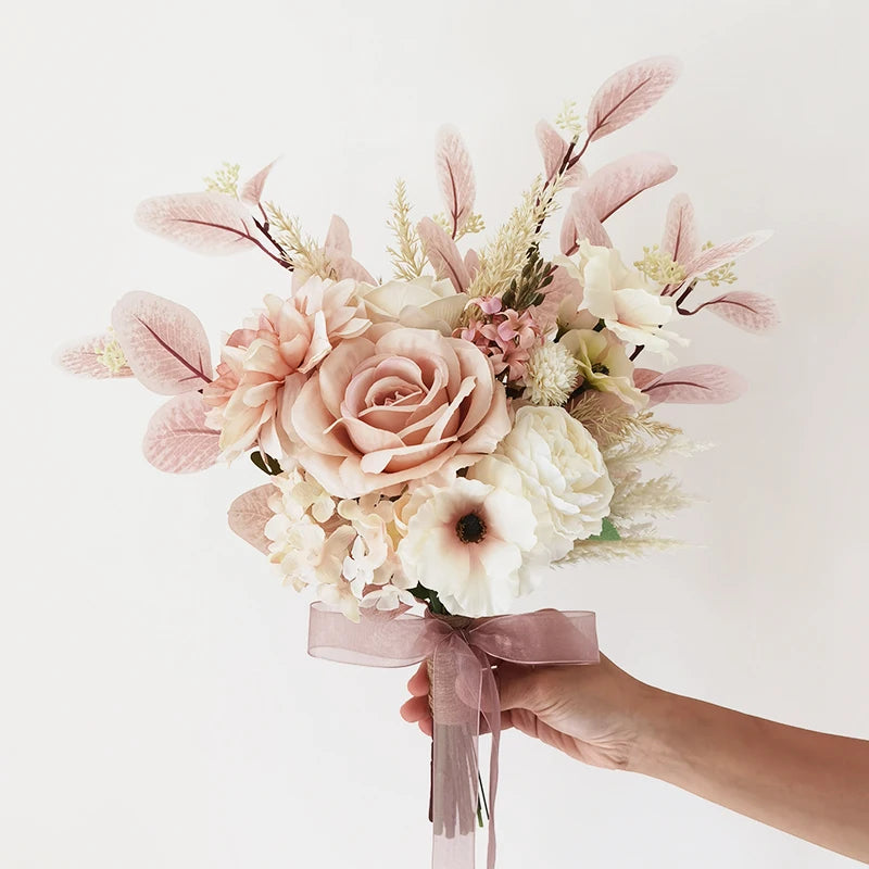 A hand holds a delicate bouquet featuring soft pink and white roses, anemones, and foliage, tied with a sheer pink ribbon against a plain white background.