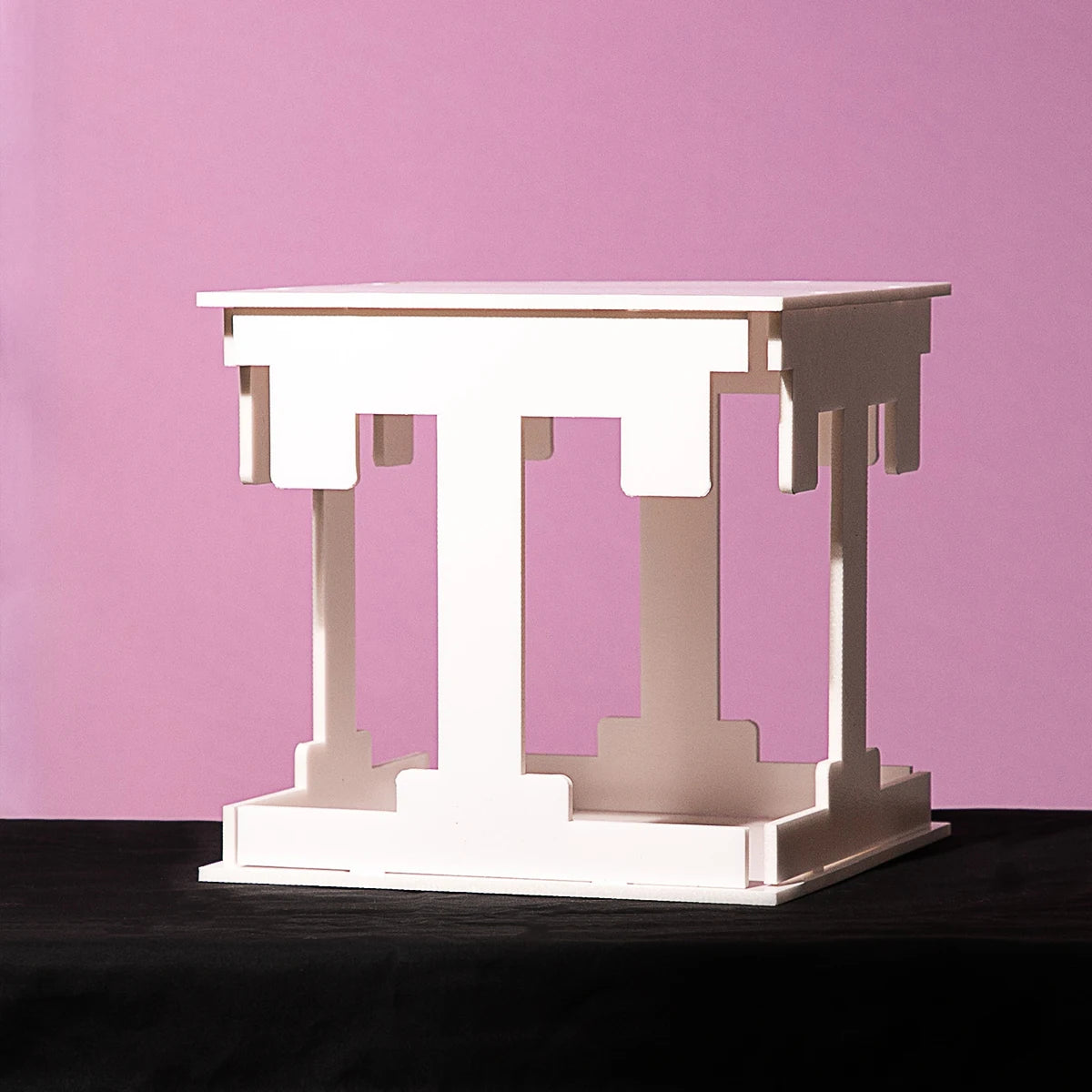 White abstract sculpture resembling a table with interlocking geometric shapes, set against a pink background and placed on a dark surface.