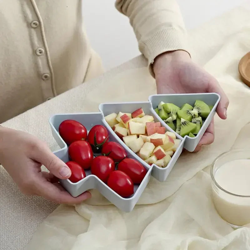 A person holds a triangular-sectioned tray containing cherry tomatoes, diced apples, and sliced kiwi. A glass of milk is nearby on a beige cloth.