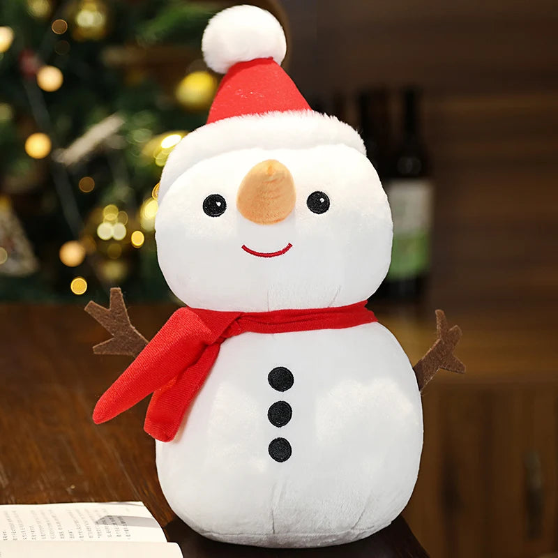 A plush snowman toy wearing a red Santa hat and scarf sits on a table, with a carrot nose and button eyes. A Christmas tree with lights is blurred in the background.