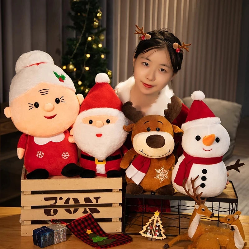 A person with reindeer antlers poses with festive plush toys of Mrs. Claus, Santa, a reindeer, and a snowman, with a Christmas tree in the background.