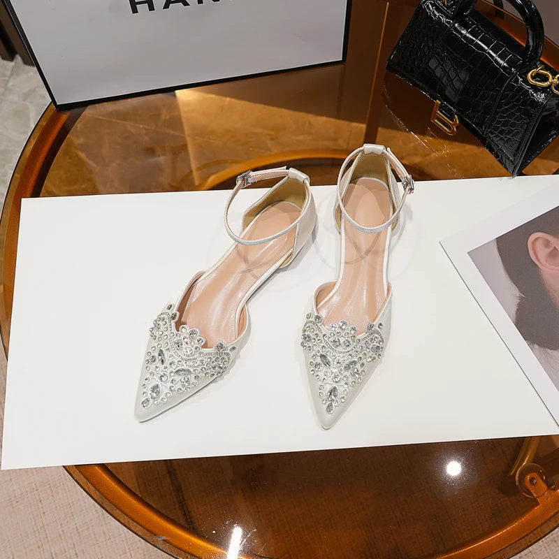 Elegant white pointed-toe heels adorned with crystals, featuring ankle straps, displayed on a white surface beside a black handbag and a fashion magazine.