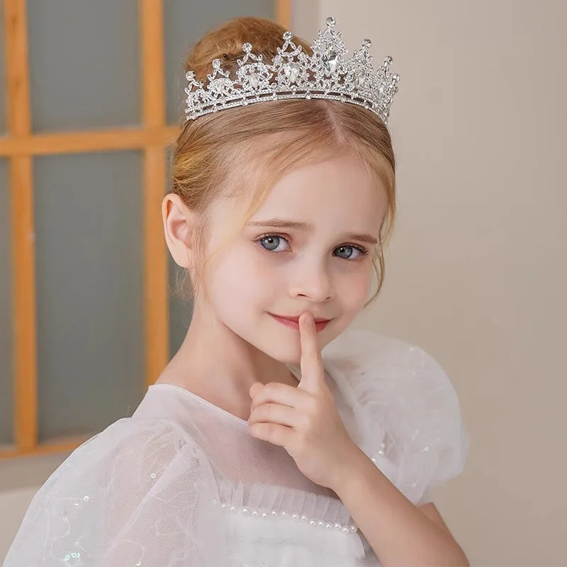 Young girl in a white dress and a jeweled tiara, smiling with a finger over her lips. Her hair is styled in an updo. The background is softly blurred with a window visible.