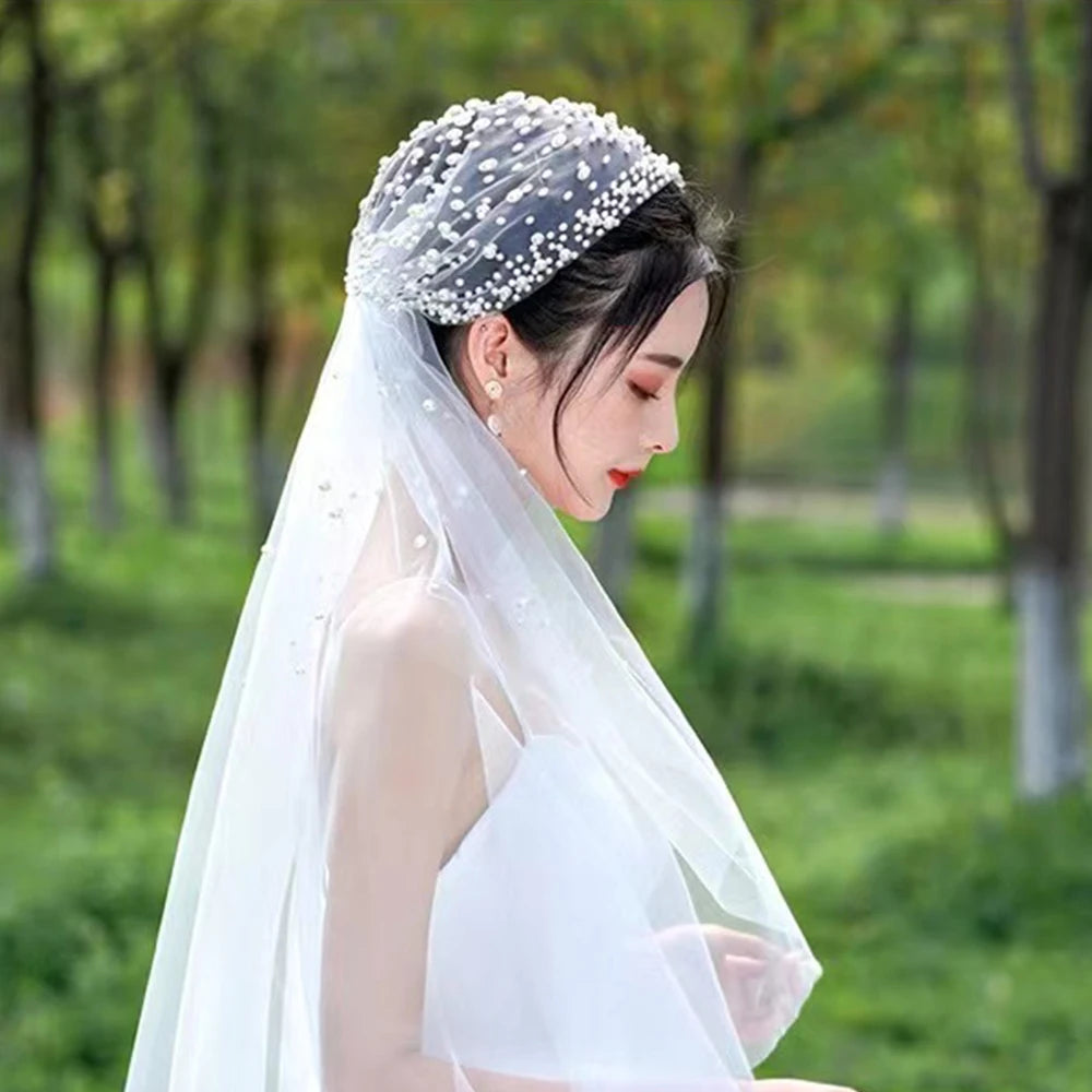 Bride in profile wearing a white dress and a pearl-embellished veil over an updo, standing outdoors with a blurred, green park background.