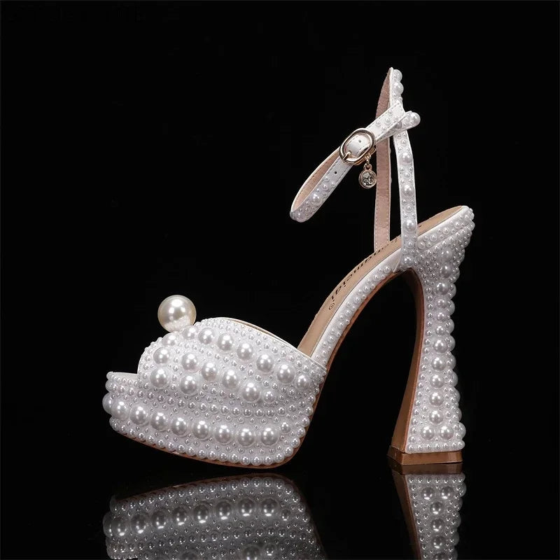 A white high-heeled shoe embellished with pearls on a black background. The shoe features a front platform, ankle strap, and a large pearl accent on the toe.