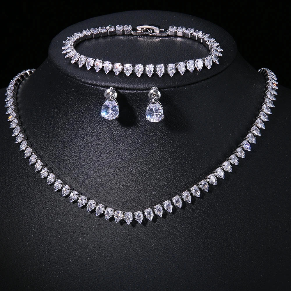 A sparkling jewelry set with a necklace, bracelet, and teardrop earrings, featuring clear diamond-like stones displayed on a black stand against a dark background.