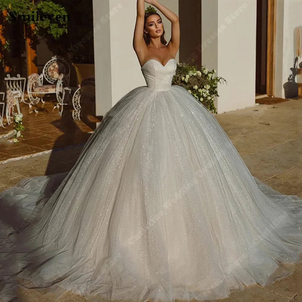 A woman in a strapless, sparkling ball gown poses elegantly outdoors, surrounded by flower arrangements and vintage-style wrought iron furniture under soft natural light.