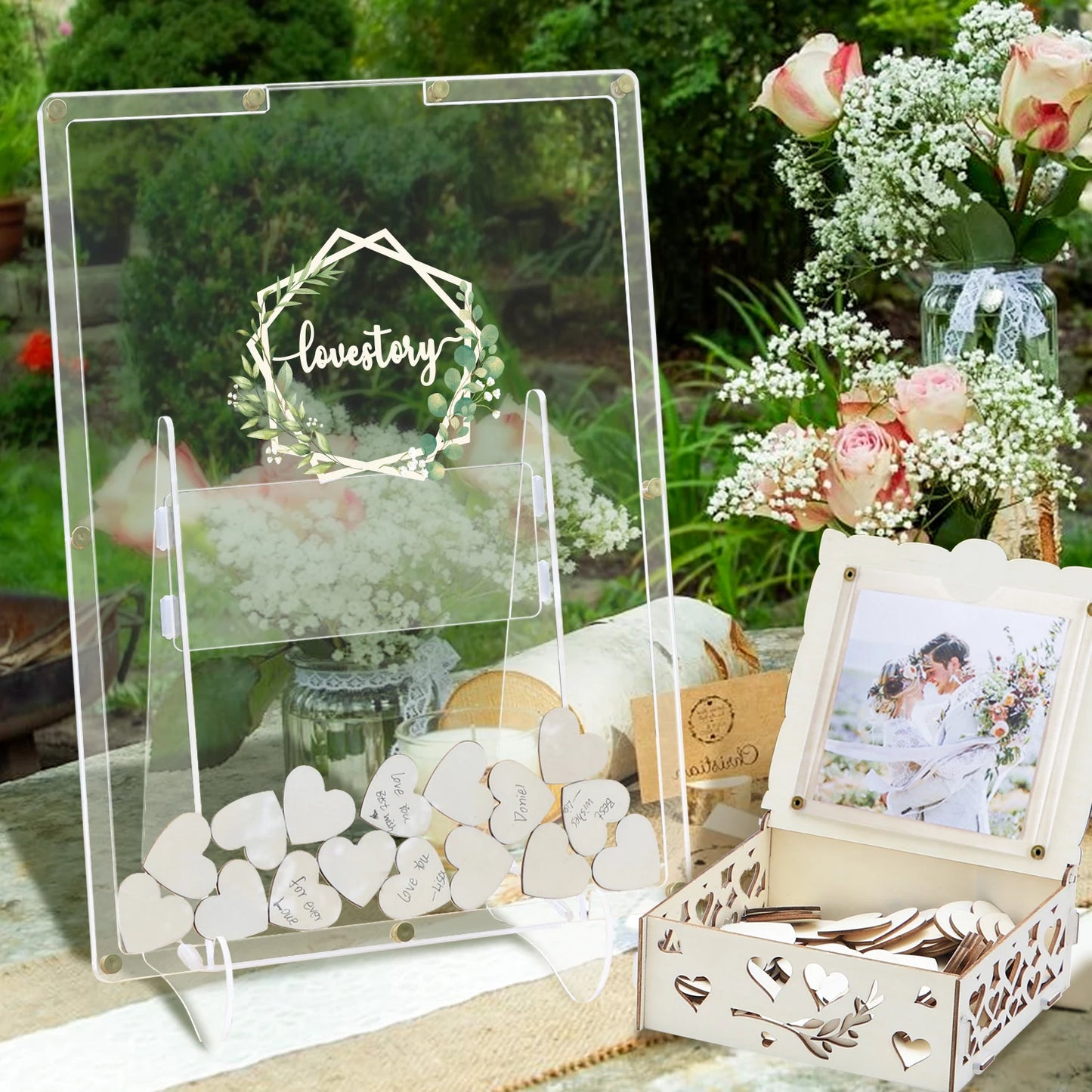A clear sign on an easel reads "lovestory," surrounded by flowers. A box with heart-shaped cards and a couple's photo sits nearby, set against a backdrop of flowers and greenery.