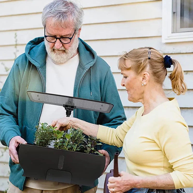 An elderly couple stands outdoors with a planter box of small plants. The man in a teal hoodie holds the planter, while the woman in a yellow top points at a plant.