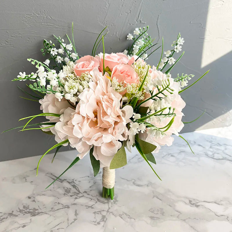 A delicate bouquet of light pink roses and peonies with white accents and greenery, wrapped in twine, rests on a marble surface against a textured gray wall.