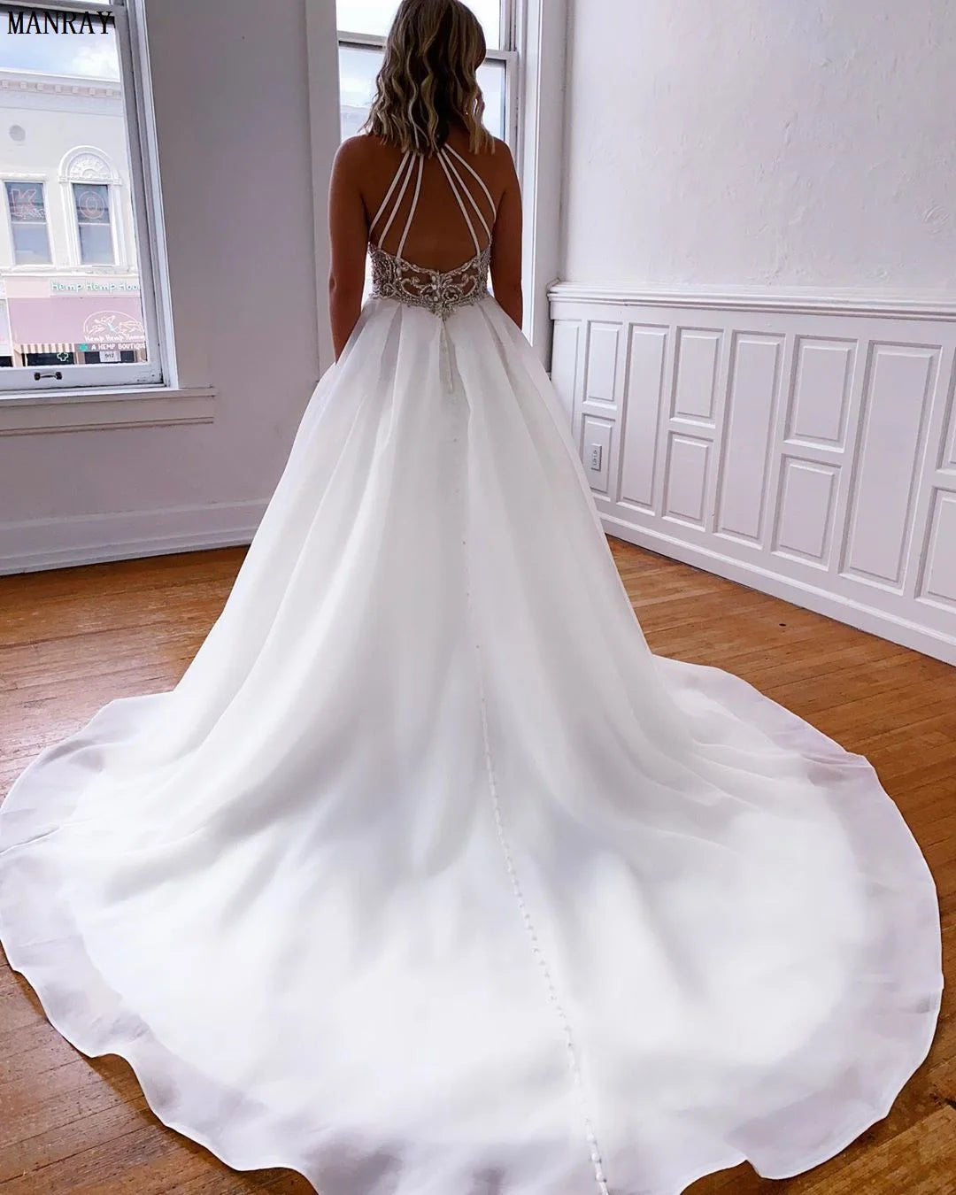 A woman in a white wedding gown with an intricate lace bodice and flowing train stands by a sunlit window, wooden floor and white paneled walls in the background.