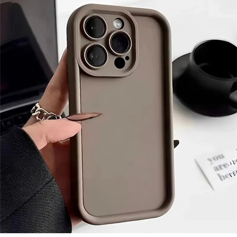 A hand holds a smartphone with a taupe case featuring a raised camera cutout. In the background, there’s a black cup and a blurred piece of paper on a white surface.