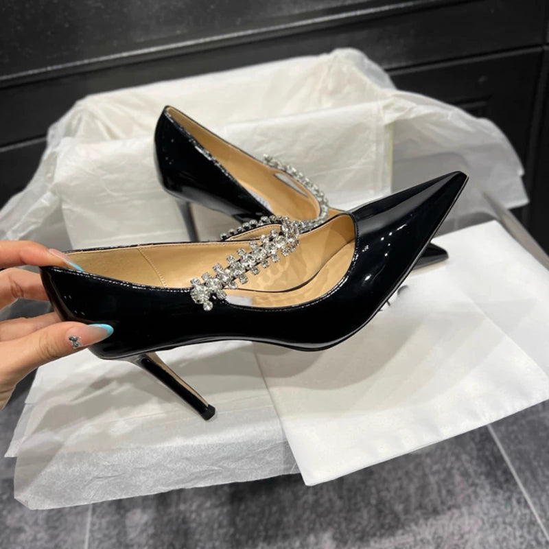 Black patent leather stiletto heels with pointed toes and crystal strap detail, placed on white tissue paper. A hand is holding one shoe for display.