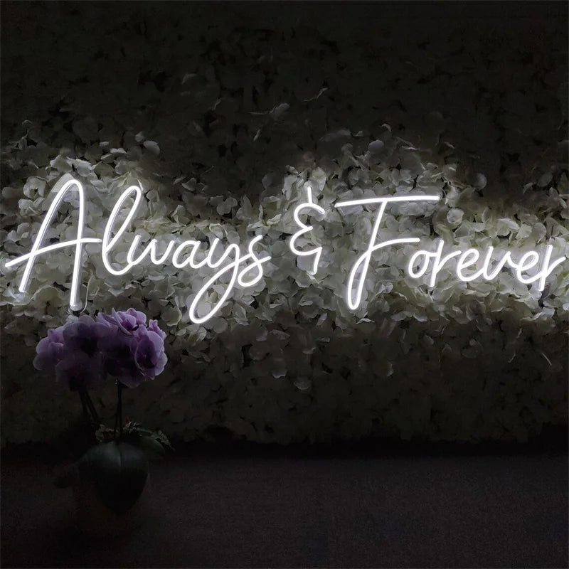 Neon sign reading "Always & Forever" in script font, illuminated on a backdrop of white flowers. Below, a small potted plant with purple blooms adds a pop of color.