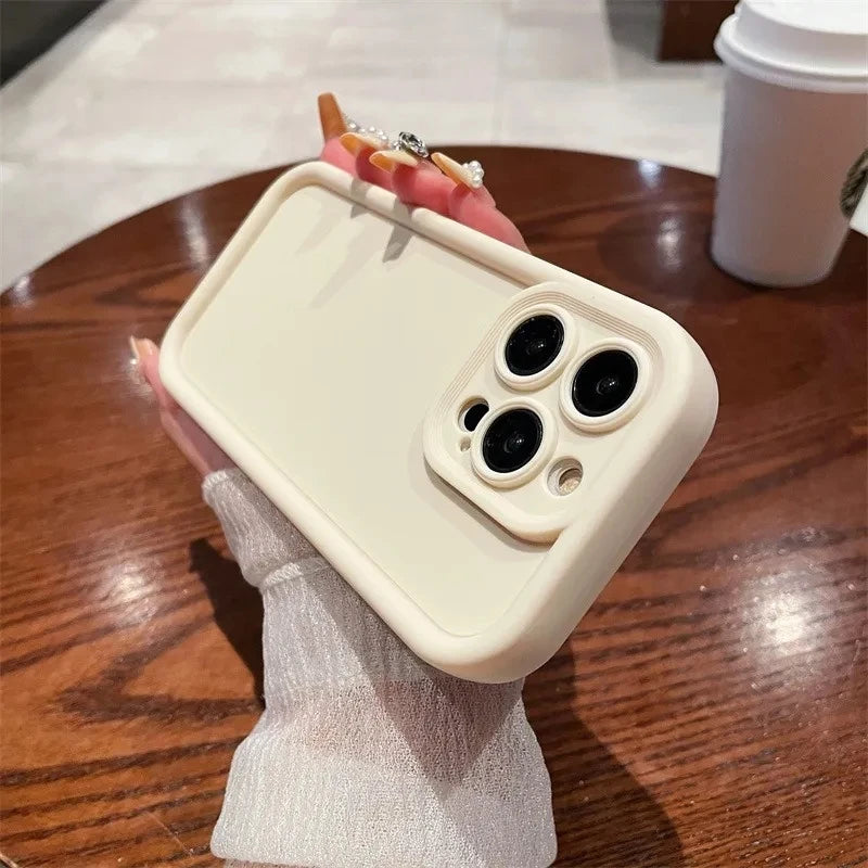 A person holds a beige smartphone with a protective case, featuring three rear cameras, over a wooden table. A takeaway coffee cup is in the background.