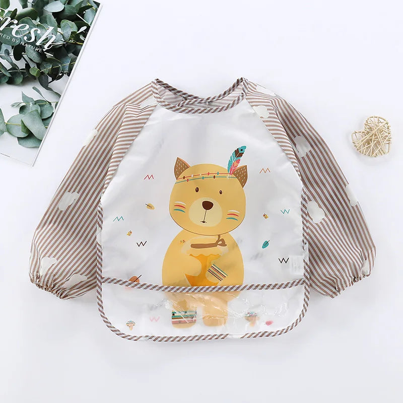 Children's bib featuring a cute bear design with striped sleeves and cloud patterns. The bear wears a feather headband and holds a honey pot. A woven heart is nearby on a smooth surface.