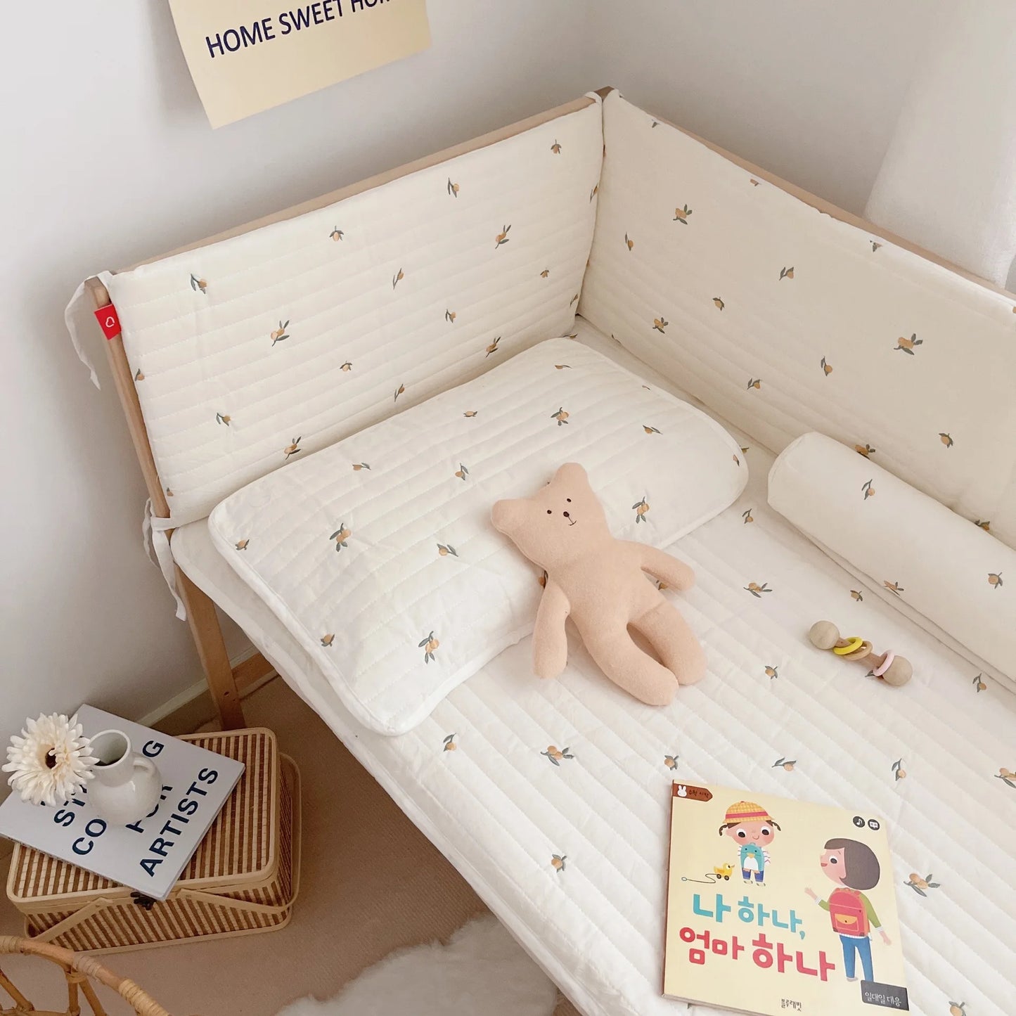 Cozy nursery with a cream quilted mattress, small floral patterns, a soft beige teddy bear, children's book, teether, and two-tier side table with books and flowers.