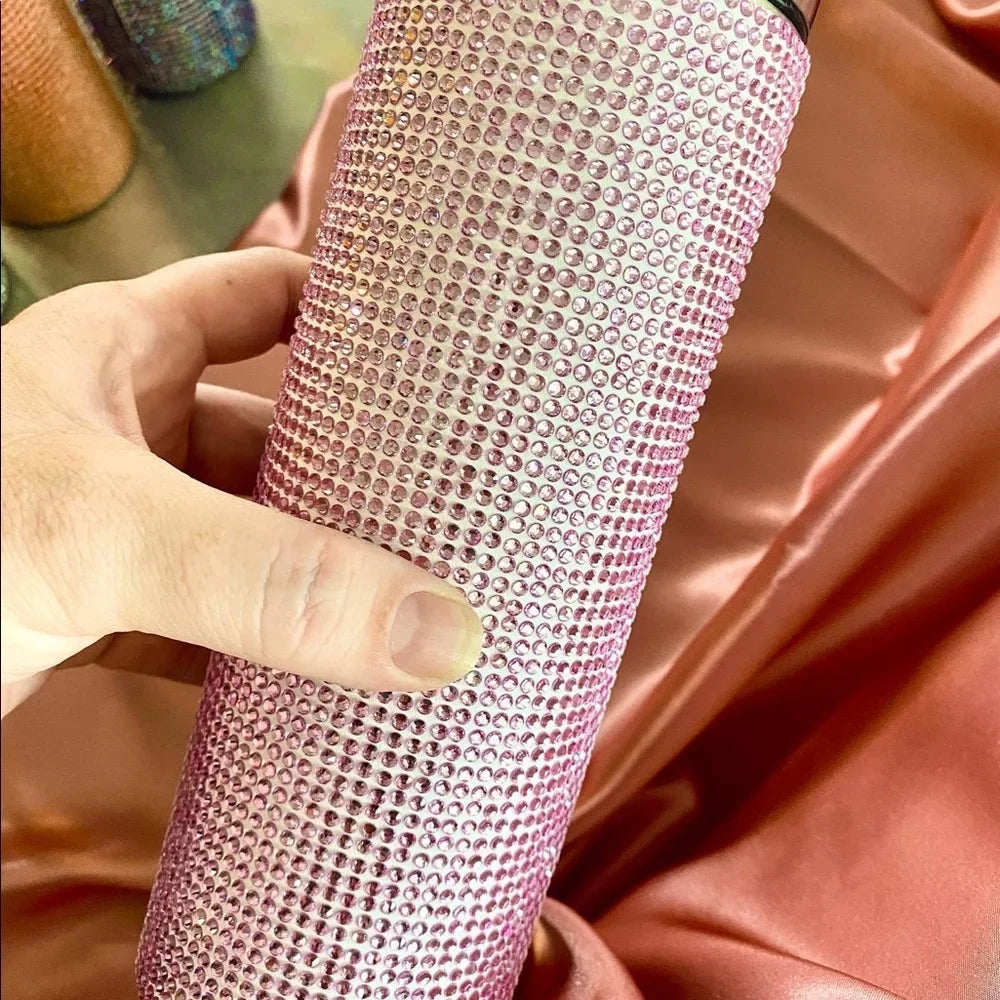 Hand holding a pink rhinestone-encrusted tumbler against a silky pink cloth. Other similarly styled items are in the background.