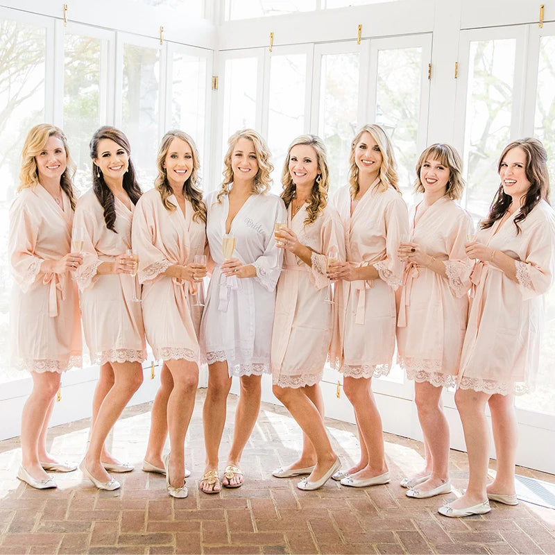 A bride in a white robe stands with seven bridesmaids in matching peach robes. They are all smiling, holding champagne glasses, in a bright room with large windows.
