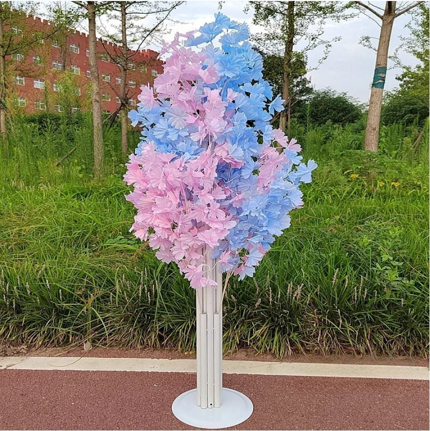 Artificial tree with pink and blue flowers in a park setting, surrounded by greenery. A red building is visible in the background under a partly cloudy sky.