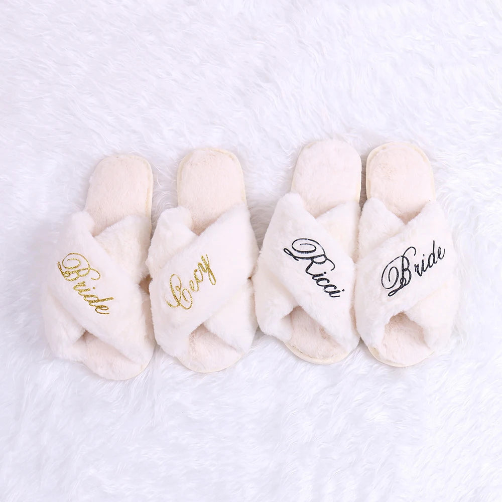 Four white fluffy slippers on a soft surface. Two pairs have "Bride" in gold and black embroidery, while the others say "Cozy" and "Ricci".