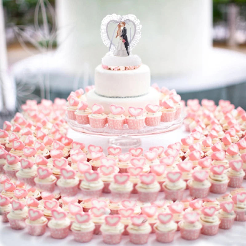 A wedding cake topped with a bride and groom figurine is surrounded by tiers of pink cupcakes with heart decorations on a white, tiered stand.