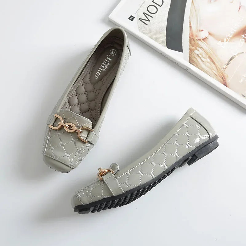 Gray quilted loafers with gold chain detail on white background, next to a fashion magazine. One shoe shows the textured sole, highlighting its design and elegance.