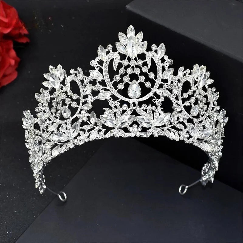 Ornate silver tiara adorned with intricate patterns and sparkling teardrop crystals, set against a dark background with a hint of red flowers on the side.