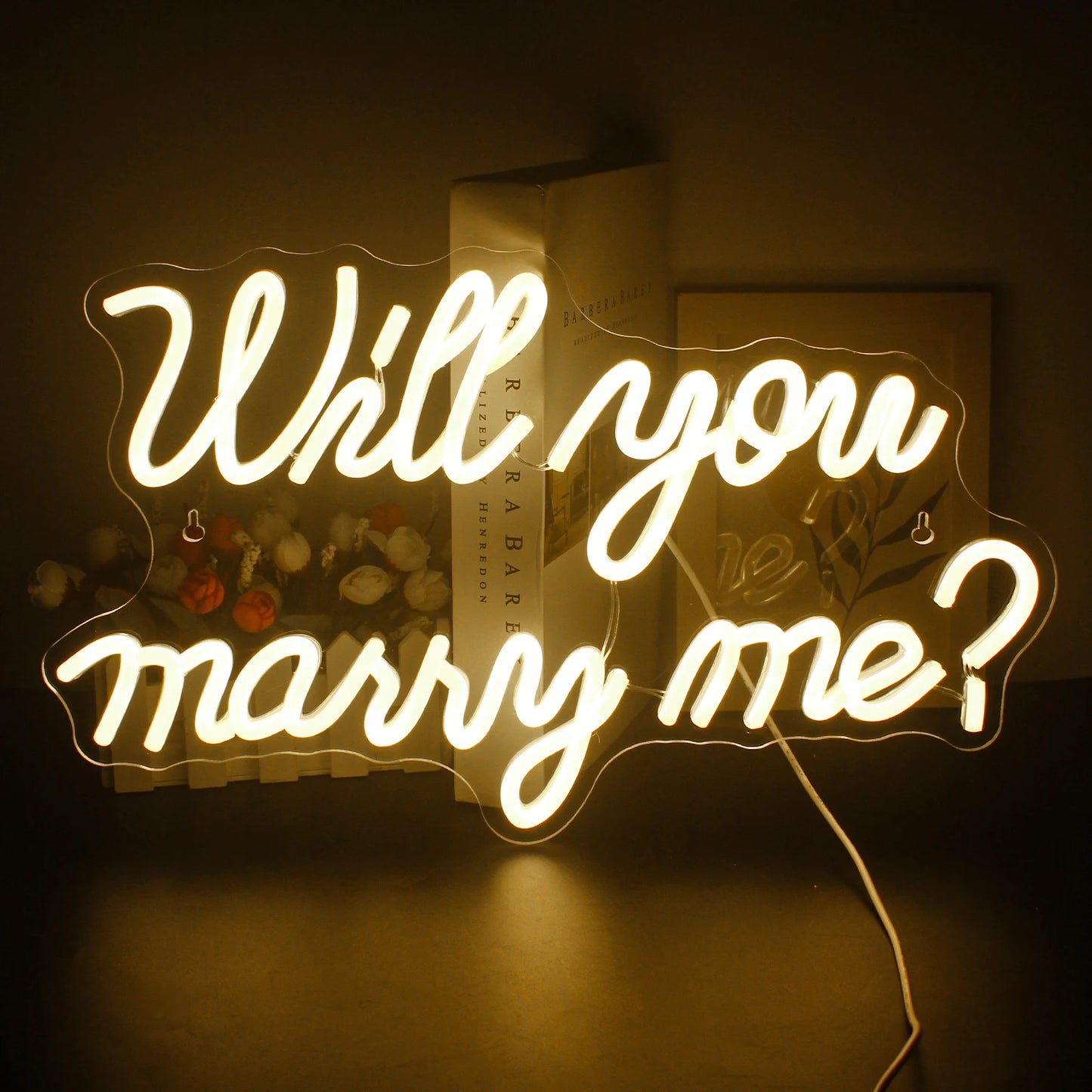 A glowing neon sign displaying the text "Will you marry me?" in script font, against a background with books and flowers, creating a warm, romantic ambiance.