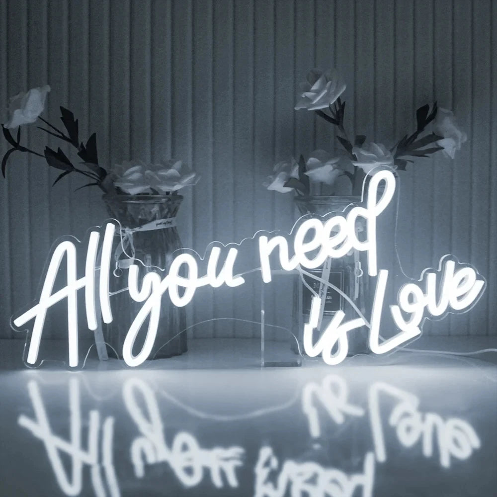 Neon sign reads "All you need is Love" with a reflection below. The background features white roses in glass vases, creating a soft, romantic ambiance.