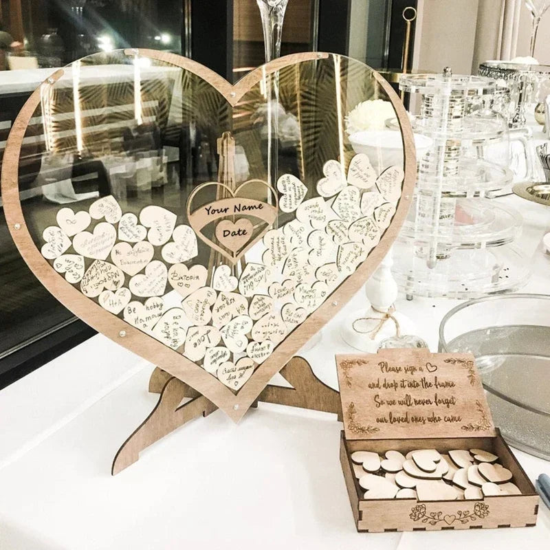 Heart-shaped guestbook with wooden hearts for messages, set atop a table with various glassware. A small box holds blank hearts awaiting signatures.