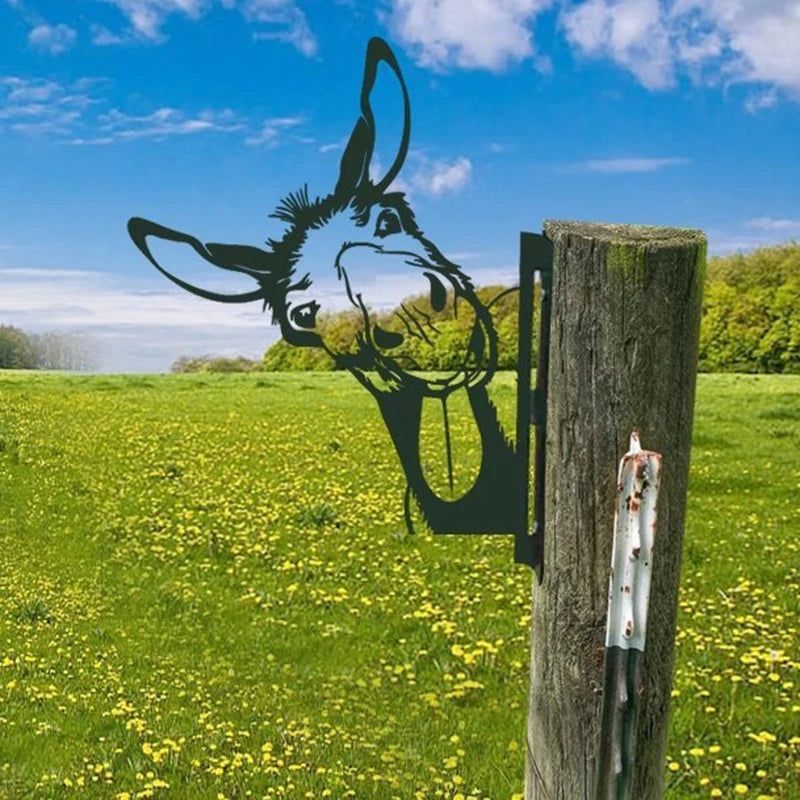 Metal silhouette of a donkey with its tongue out mounted on a wooden post, set against a lush green field with yellow flowers and a clear blue sky.
