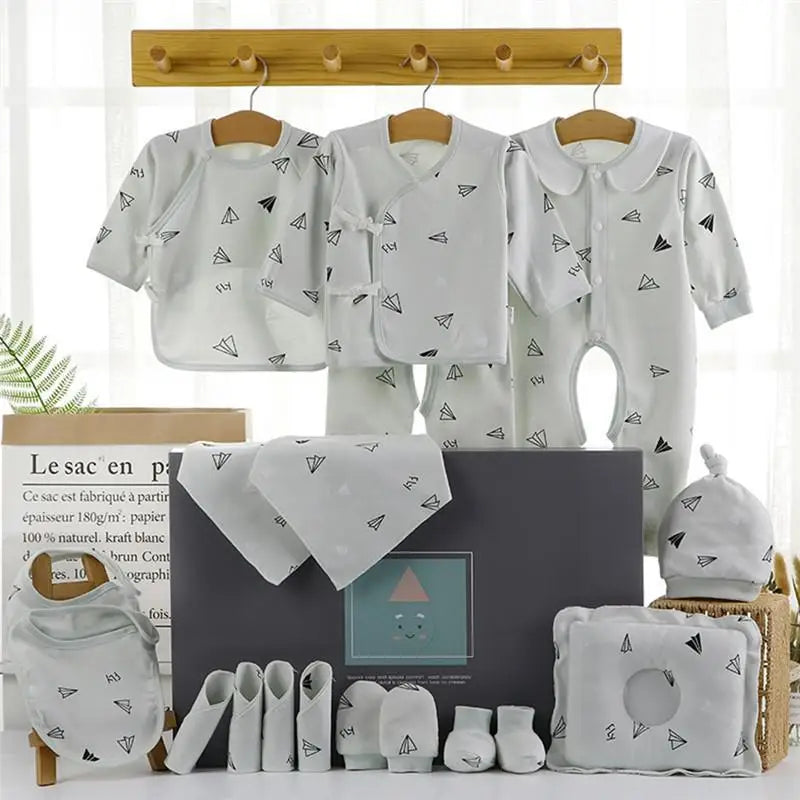Baby clothing set in white with black paper airplane pattern. Includes onesies, bib, hat, mittens, socks, pants, and washcloths arranged with a decorative box on a shelf.