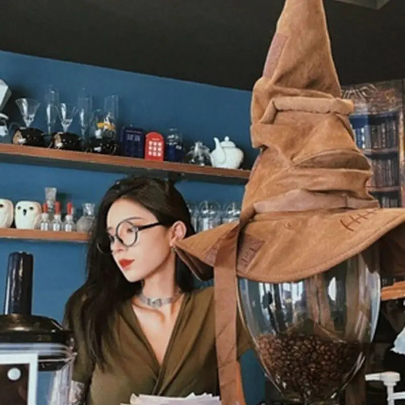 Woman in glasses sits near a brown, pointed hat atop a glass jar filled with coffee beans. Shelves with teapots and cups adorn the blue wall behind her.
