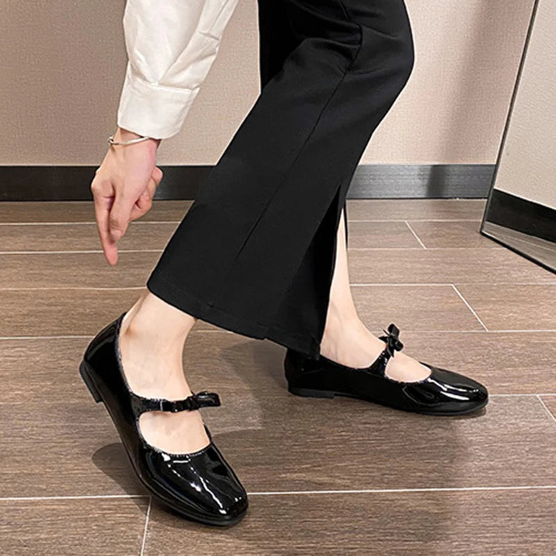 Person adjusting shiny black Mary Jane shoes with bows, wearing black pants and a white shirt. The floor is wooden, and there is a mirror in the background.