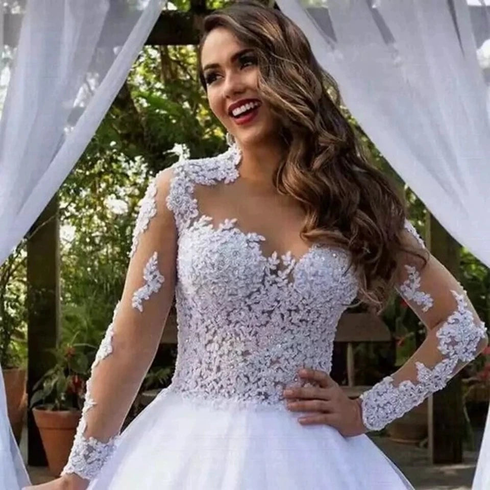 Woman in a detailed white lace wedding gown with sheer sleeves, smiling by an outdoor pergola draped with white fabric, surrounded by greenery.