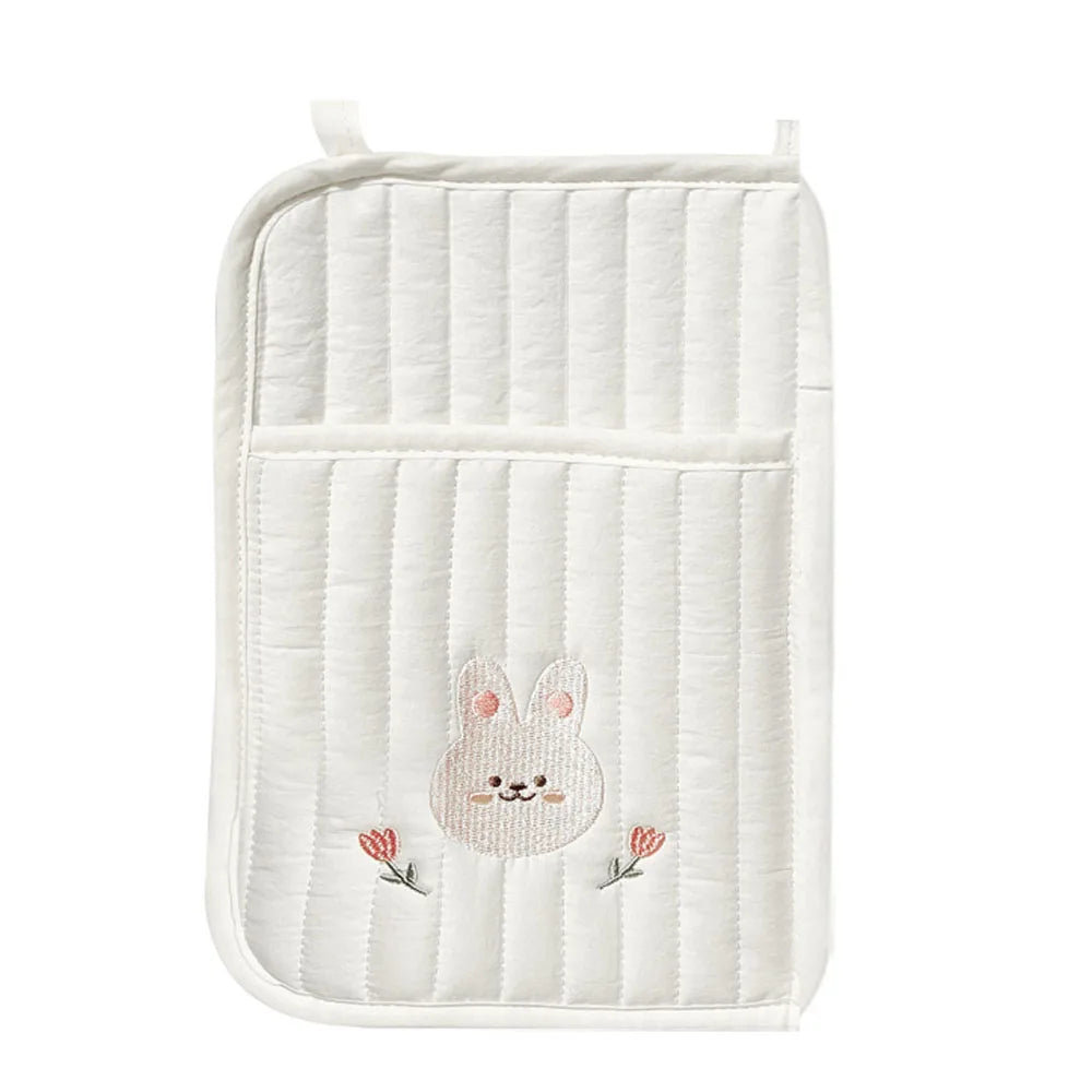 White quilted fabric with vertical stitching, featuring an embroidered bunny face with pink cheeks. Two small embroidered tulips are below the bunny. A loop is at the top.