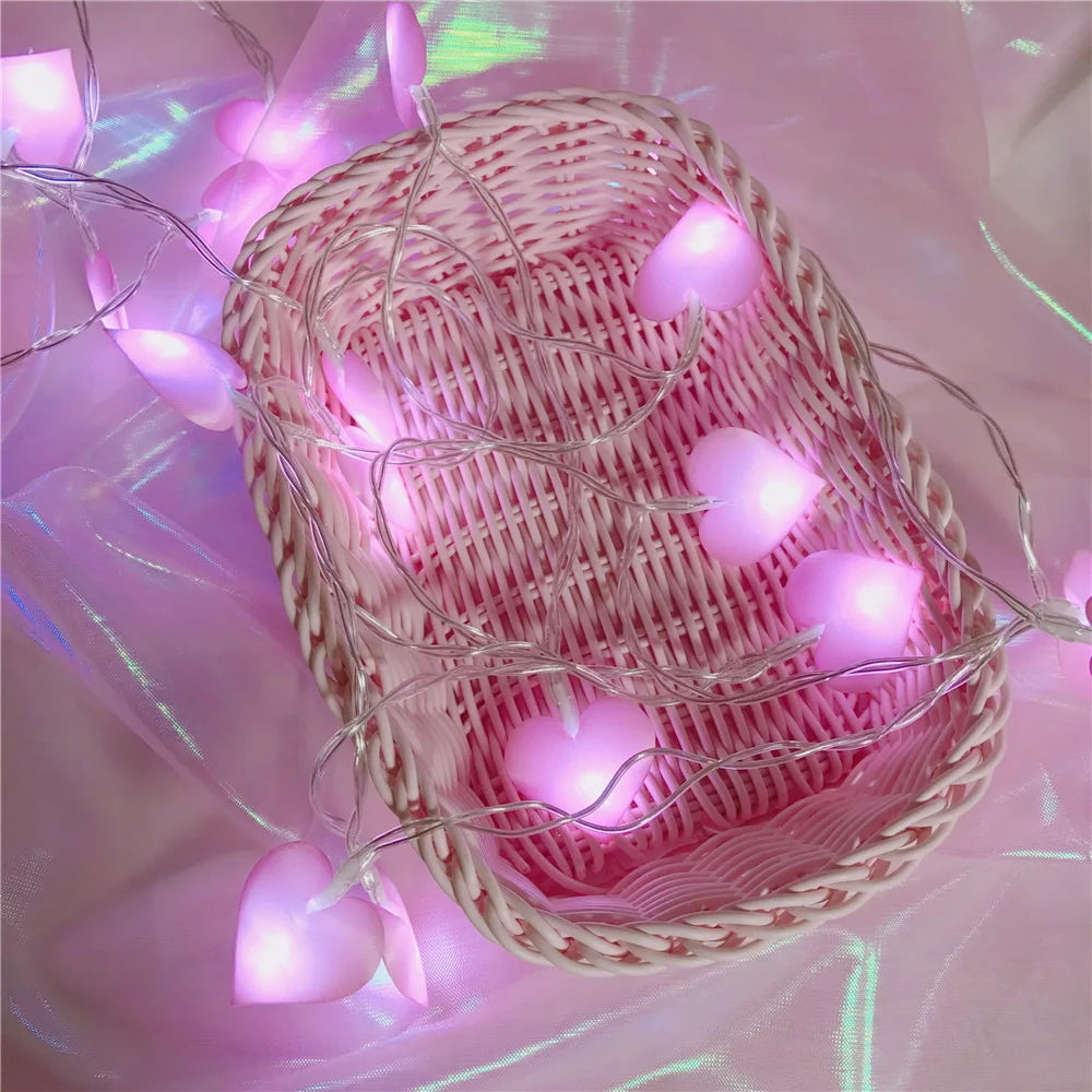 Pink heart-shaped lights draped over a pink wicker basket on a shiny fabric background. The lights cast a soft, ambient glow, enhancing the dreamy aesthetic.
