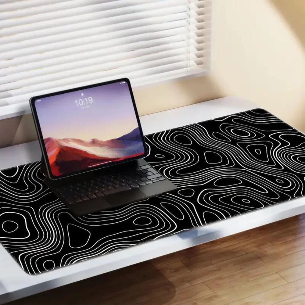 Tablet with keyboard on a desk covered by a black mat with abstract white contour lines, near a window with blinds partially open, letting in natural light.