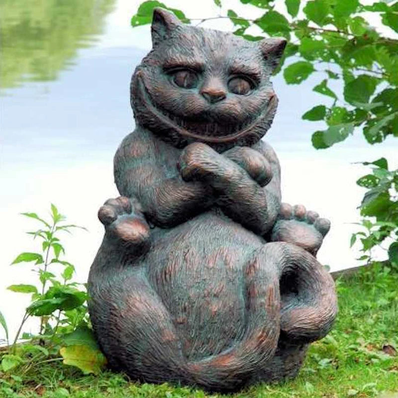 Bronze statue of a grinning, seated cat with crossed paws, wide eyes, and curled tail, set amidst green foliage by a calm lakeside.