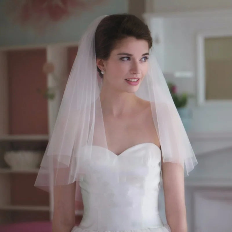 Bride in a strapless white gown, wearing a delicate tulle veil, stands indoors with a gentle smile. Background softly blurred, highlighting her elegant appearance.