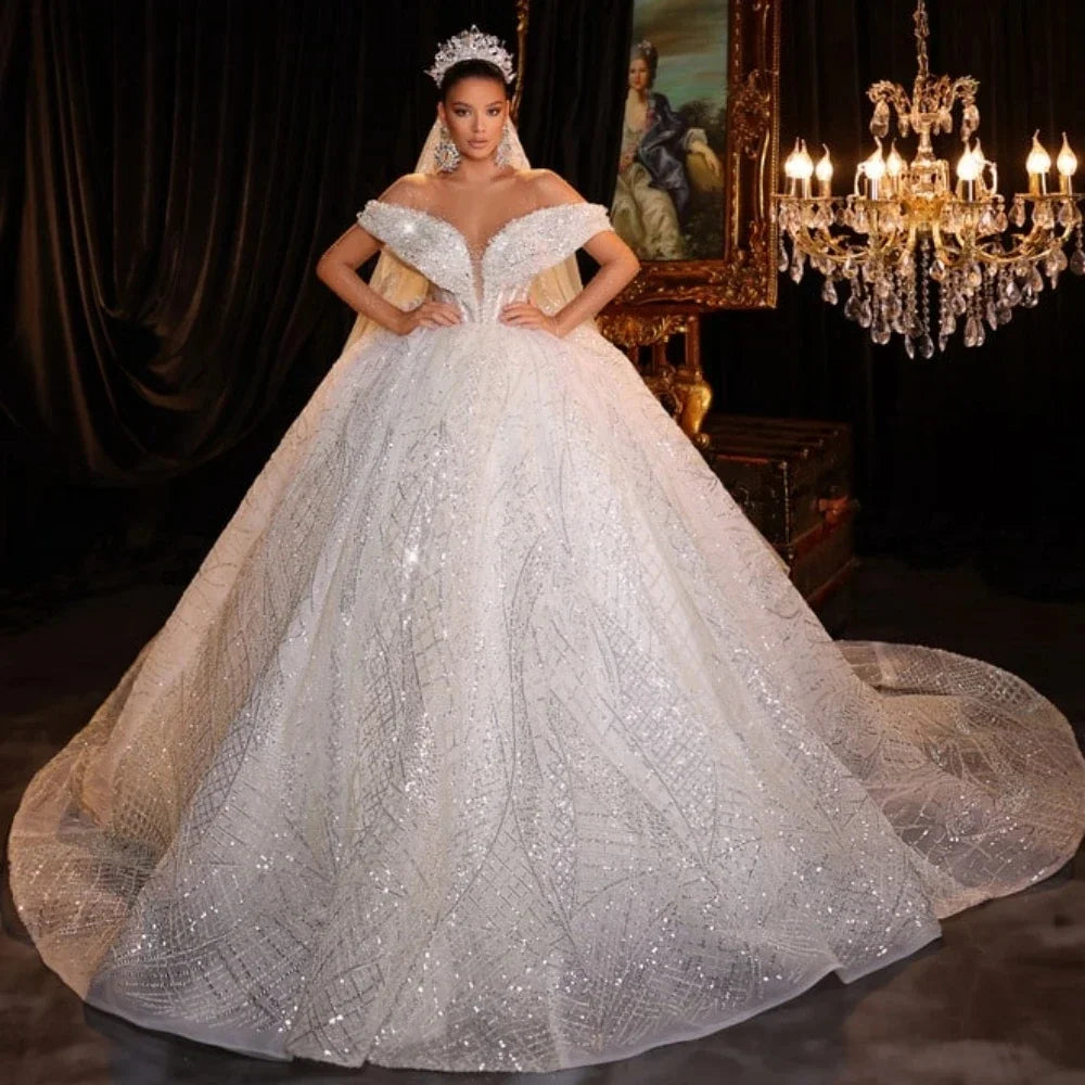 A woman in a sparkling, off-shoulder, ball gown-style wedding dress with a long train stands confidently under a chandelier, surrounded by elegant decor and a painting.