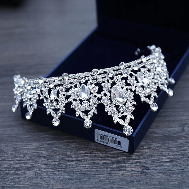 A sparkling silver tiara encrusted with large teardrop crystals and intricate floral details is displayed on a dark velvet box, set against a wooden surface.