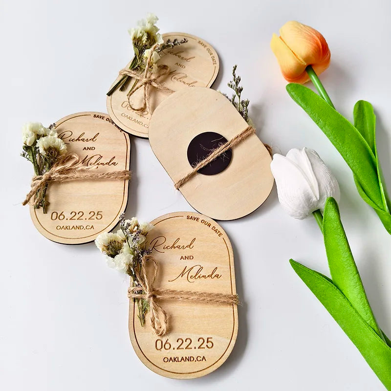 Wooden save-the-date magnets with names, date, and location are tied with twine and small flowers. Nearby, there are orange and white tulips on a white surface.